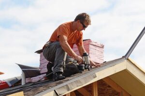 A roofer stands atop a home installing gray asphalt shingles with nearby rolls of roofing material visible.